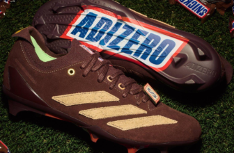 Snickers x adidas Adizero Electric+ II football cleats with the Snickers wrapper print on the outsole, shown on grass with Snickers bars